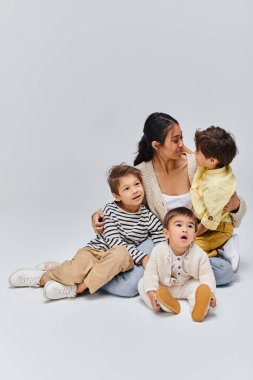 Genç bir Asyalı anne, çocuklarıyla birlikte gri arka planda sakin bir stüdyoda yerde oturuyor..