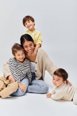 Genç bir Asyalı anne yerde oturuyor, sakin ve sevgi dolu bir anda çocuklarını şefkatle kucaklıyor..