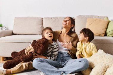 İki çocuğu ve oyuncak ayısıyla yerde oturan genç bir Asyalı anne sıcak bir aile anı yaratıyor..