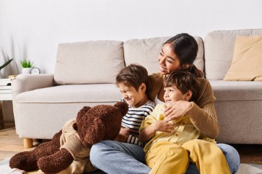 Genç bir Asyalı anne yerde iki çocuğu ve bir oyuncak ayıyla oturuyor. İçtenlik ve sevgiyle çevrili..