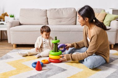 Genç bir Asyalı anne, küçük oğluyla neşeyle etkileşime geçer. Evlerinin oturma odasında yerde birlikte oynarlar..