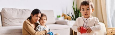 A young Asian mother and her two little sons sit closely together on a cozy couch in their living room. clipart