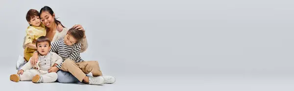 stock image A young Asian mother sitting on the ground with her children, engaging in a moment of togetherness in a studio on a grey background.