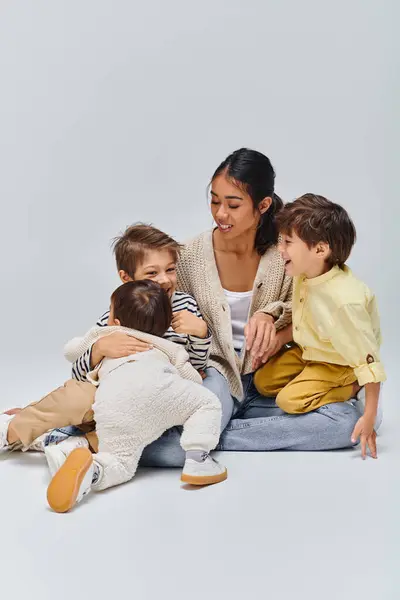 Asian mother sits on floor embracing children in studio against grey backdrop.