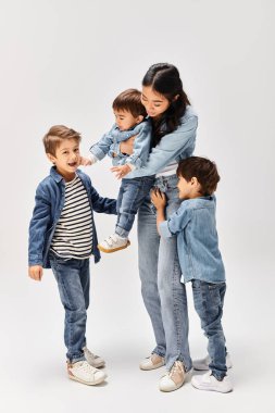 Aralarında Asyalı bir anne ve küçük oğullarının da bulunduğu bir grup çocuk, gri bir stüdyoda yan yana duruyorlar, hepsi kot elbise giyiyorlar..
