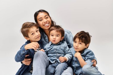 A young Asian mother wearing denim sits with children, her little sons also in denim, in a grey studio setting. clipart