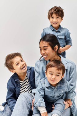 Bir grup genç çocuk, bir anne ve oğulları, hepsi kot giyip gri bir stüdyoda yan yana oturuyorlar..