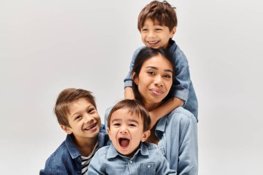 Aralarında genç bir Asyalı anne ve kot elbiseli küçük oğullarının da bulunduğu bir grup çocuk gri bir stüdyoda fotoğraf için poz veriyorlar..