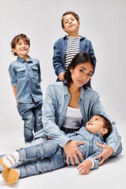 Genç bir Asyalı anne, gri bir stüdyoda, kot giyip oğullarıyla birlikte yerde oturuyor..