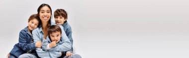 A young Asian mother in denim sits atop her young sons also wearing denim in a grey studio setting. clipart