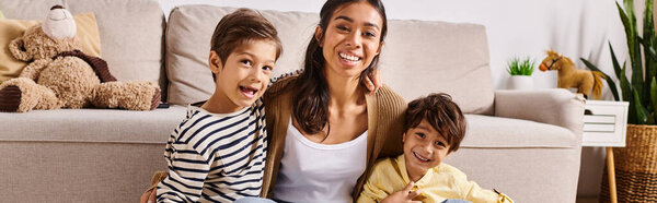 A young Asian mother and her two little sons sitting together on a couch in their homes living room, sharing a moment of bonding and connection.