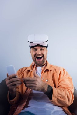 Sandalyede oturan adam, telefon ekranına dalmış, bir stüdyo ortamında sanal gerçeklik teknolojisiyle çevrili..