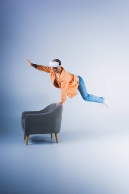 A man in a VR headset performs a gravity-defying trick on a chair in a futuristic studio setting. clipart