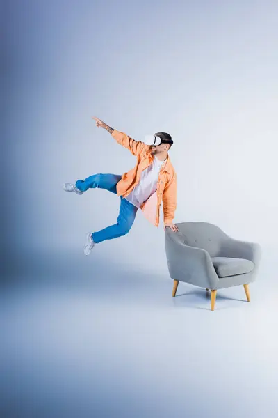 stock image A man wearing a VR headset jumps energetically in a studio, soaring over a chair with agility and grace.