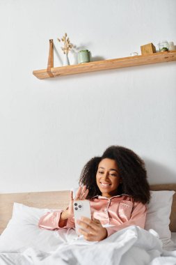 A curly African American woman in pajamas takes a selfie with her phone while laying in bed in the morning. clipart