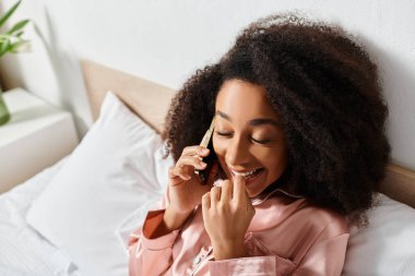 Pijamalı kıvırcık Afrikalı Amerikalı kadın sabahları yatakta uzanırken cep telefonuyla konuşuyor..