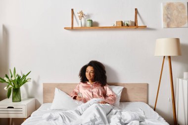 Pijamalı kıvırcık Afrikalı Amerikalı bir kadın sakin bir sabahta, rahat bir yatak odasında beyaz çarşaflı bir yatakta huzur içinde oturur..