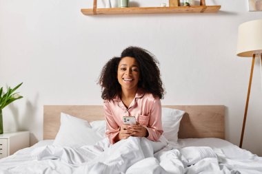 Pijamalı kıvırcık Afrikalı Amerikalı kadın, yatakta oturuyor, cep telefonuna dalmış. Sabah ışığı pencereden süzülüyor.