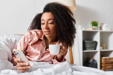 Kıvırcık Afrikalı Amerikalı kadın pijamalarıyla sabah kahvesinin tadını çıkarıyor ve yatakta dinlenirken cep telefonunu kontrol ediyor..