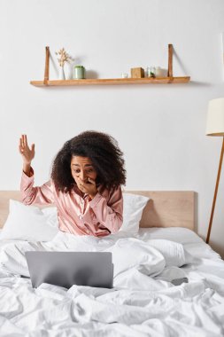 Kıvırcık Afrikalı Amerikalı bir kadın yatakta oturuyor. Sabahları rahat yatak odasında dizüstü bilgisayar kullanıyor..