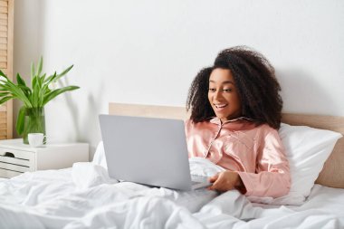 Yatakta pijamalı Afro-Amerikalı bir kadın sabahları yatak odasında dizüstü bilgisayar kullanıyor..