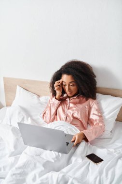 Pijamalı kıvırcık Afrikalı Amerikalı kadın yatakta oturmuş, telefonla konuşuyor..