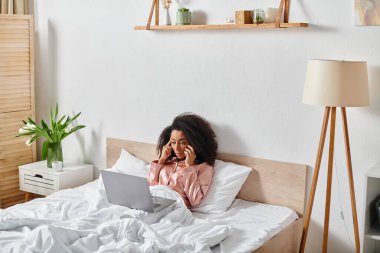 Yatakta pijamalı kıvırcık Afrikalı Amerikalı bir kadının dizüstü bilgisayarını kullanmaya odaklandığı sakin bir sabah sahnesi..