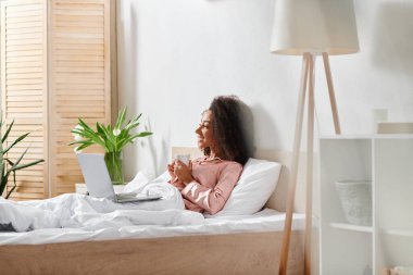 Yatakta pijamalı kıvırcık Afrikalı Amerikalı kadın, dizüstü bilgisayara gömülmüş, sabah ışığı pencereden süzülüyor..
