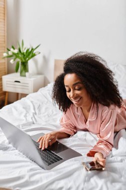 Sabahları rahat bir yatak odasında dizüstü bilgisayar kullanan kıvırcık saçlı, pijamalı Amerikalı bir kadın..