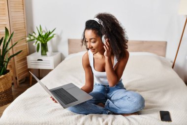 Askılı tişörtlü kıvırcık Afrikalı Amerikalı kadın modern yatak odasında dizüstü bilgisayar kullanarak yatakta oturuyor..