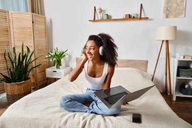 Curly African American woman in a tank top sitting on a bed, using a laptop and wearing headphones in a modern bedroom. clipart