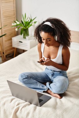 Kolsuz tişörtlü kıvırcık Afrikalı Amerikalı kadın, modern yatak odasında cep telefonu ekranı tarafından emilmiş bir şekilde yatakta oturuyor..