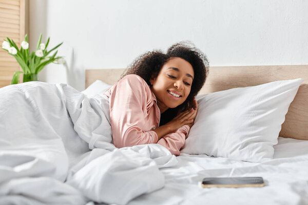 Curly African American woman in pajamas peacefully reclines on a bed with white sheets in a cozy bedroom.
