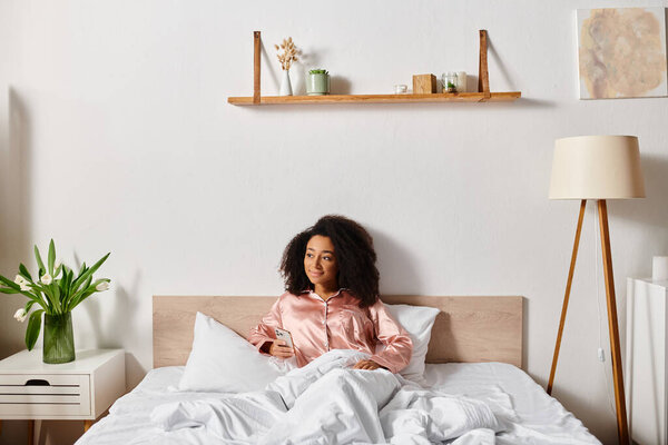 A curly African American woman in pajamas sits peacefully on a bed with white sheets in a cozy bedroom on a calm morning.