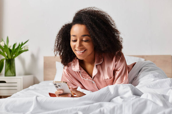 A curly African American woman in pajamas lays in bed, absorbed in her cell phone screen, surrounded by warm morning light.