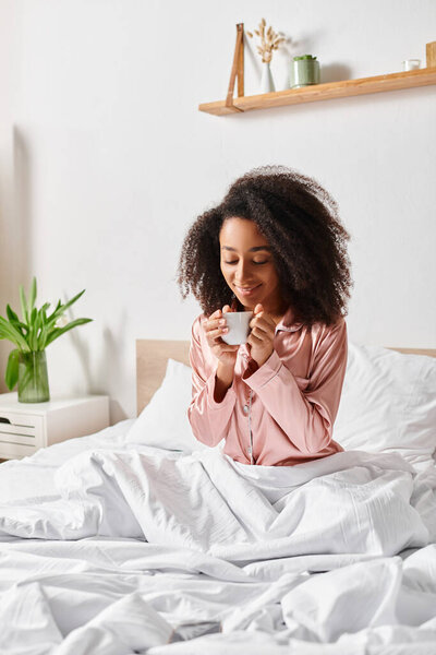 A curly African American woman in pajamas sitting on a bed, peacefully sipping a cup of coffee in the morning.