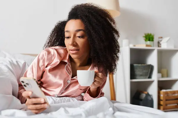 Curly Afrikansk Amerikansk Kvinne Pyjamas Nyter Morgenkaffe Sjekke Hennes Mobiltelefon – stockfoto