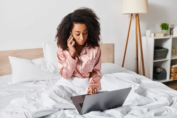 Curly African American woman in pajamas sits on bed, focused on using laptop in cozy bedroom during morning time.
