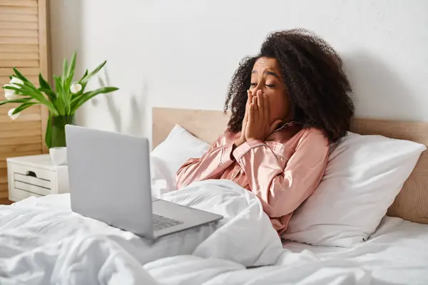stock image A curly African American woman in pajamas lies in bed with a laptop on her lap, engaging with technology in a cozy bedroom setting.