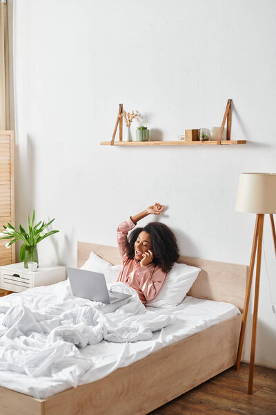 A curly African American woman in pajamas, engrossed in her laptop, lays on a cozy bed in the morning light.