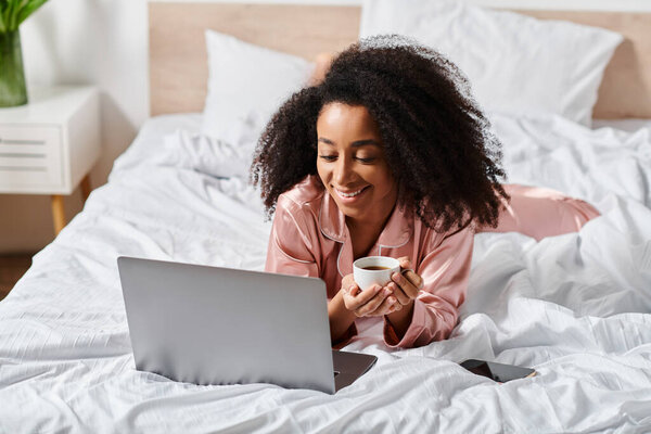 Curly African American woman in pajamas enjoys a peaceful morning in bed with a laptop and a cup of coffee.
