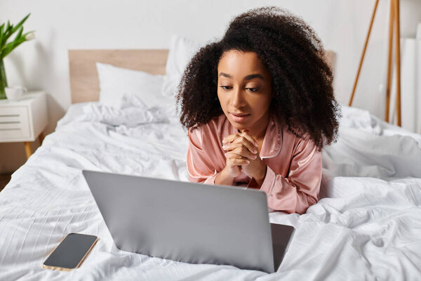 A curly African American woman in pajamas lays on a bed, peacefully using a laptop in a cozy bedroom in the morning.