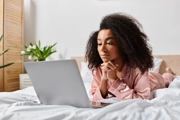 stock image Curly African American woman in pajamas lying in bed, absorbed in using a laptop computer.