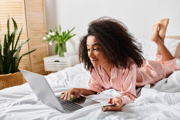 A curly African American woman in pajamas lays on a bed, focused on her laptop screen in a cozy bedroom during morning.