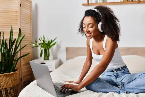 Curly African American woman in a tank top, sitting on a bed, intensely focused on using a laptop computer in a modern bedroom.