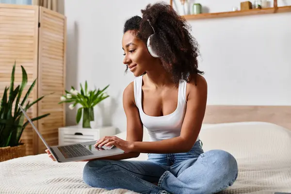 ベッドの上に座っているタンクの上のカーリーアフリカ系アメリカ人女性は 現代のベッドルームでラップトップコンピュータを使用して光沢 — ストック写真