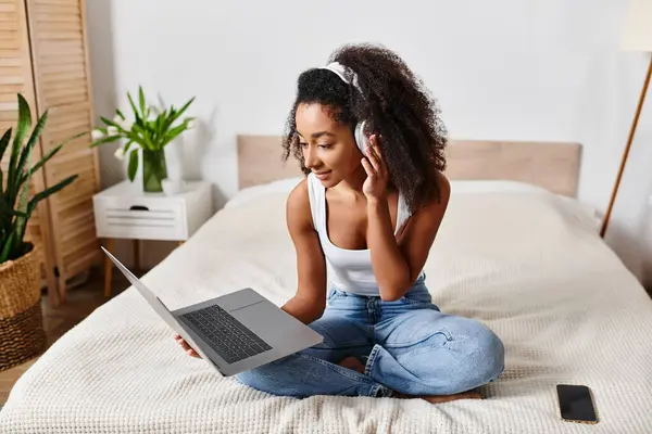 Curly African American woman in tank top sits on bed using laptop computer in stylish modern bedroom.