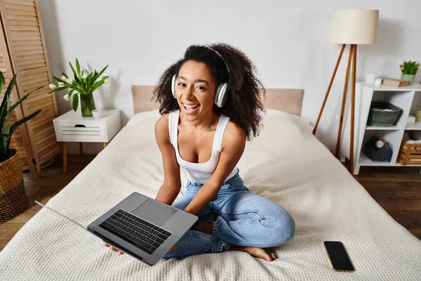 A curly African American woman in a tank top sits on a bed with a laptop and headphones, engrossed in her digital activities.