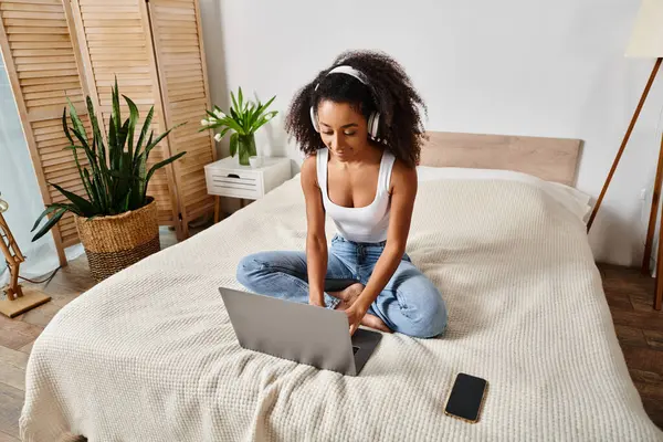 タンクトップのカーリーアフリカ系アメリカ人女性がベッドの上に座り 現代の寝室でラップトップコンピュータを使用して光沢 — ストック写真