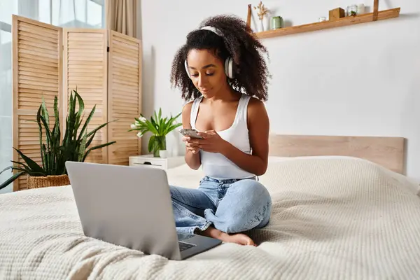 Curly African American woman sitting on bed, engrossed in laptop screen, in a modern bedroom.